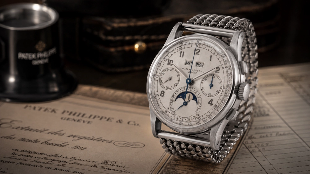 Yakın planda, pirinç tanesi örgülü çelik bileziğe, gümüş rengi kadrana, ay evresi göstergesine, kronograf ve sonsuz takvim fonksiyonlarına sahip, Patek Philippe logolu büyüteç ve eski kayıt belgeleri üzerinde zarifçe duran, ikonik Patek Philippe Ref. 1518 çelik kol saati görülüyor.