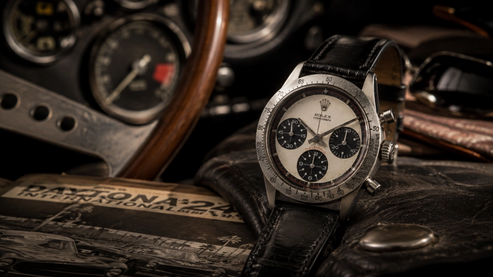 Siyah deri kayışlı, beyaz kadranlı ve siyah alt kadranlı ikonik bir Rolex Daytona Paul Newman kronograf saati, eski bir spor arabanın ahşap direksiyonu ve göstergelerinin bulanık arkaplanında, Daytona International yazılı bir dergi ve yıpranmış deri bir yüzey üzerinde konumlanmış.