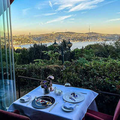 İstanbul Boğazı'nın eşsiz manzarasını sunan bir terasta, gün batımının turuncu ve mor renkleriyle aydınlanan gökyüzü altında, iki kişilik şık bir masanın özenle hazırlandığı ve bu ortamın İstanbul'un Michelin Yıldızlı restoranlarının sunduğu unutulmaz yemek deneyimini gözler önüne serdiği etkileyici bir kare.