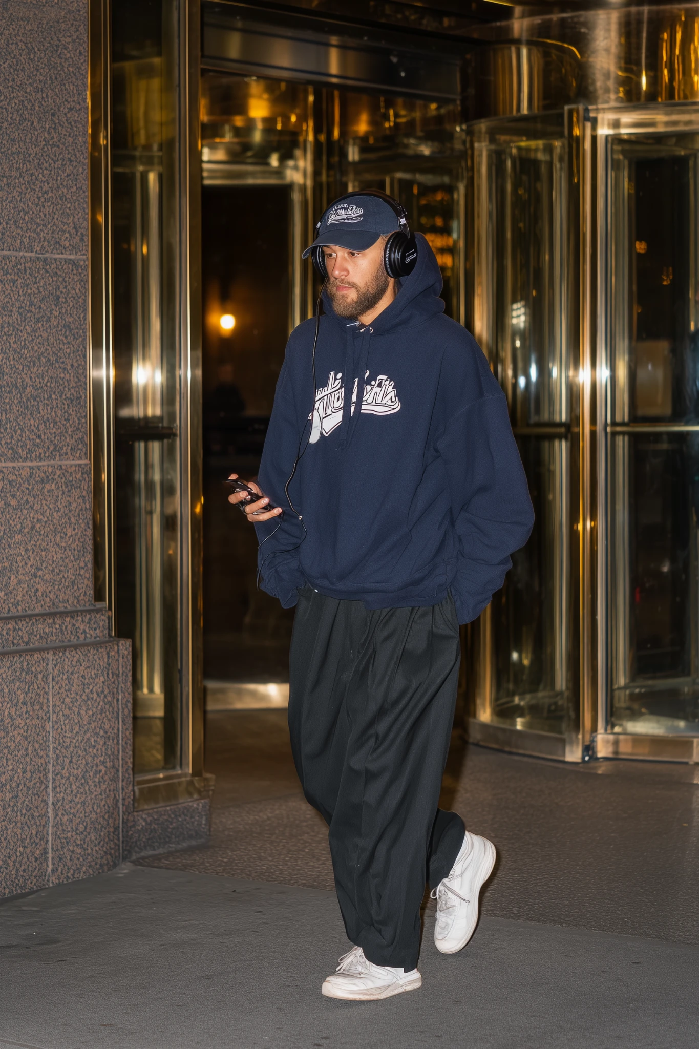 Gece şehir ışıkları önünde kulaklık takan erkek; lacivert hoodie, rahat pantolon ve sneaker ile şehirli sokak stili.