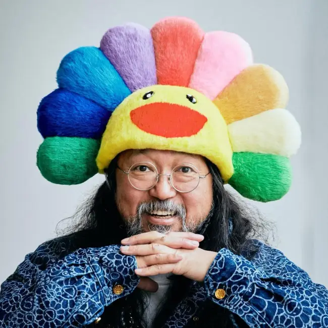 Takashi Murakami, ikonikleşmiş renkli çiçek şapkasını takarken gülümsüyor. Japon sanatçı, neşeli ve pop-art estetiğini yansıtan tarzıyla küresel sanat sahnesinde öne çıkıyor.