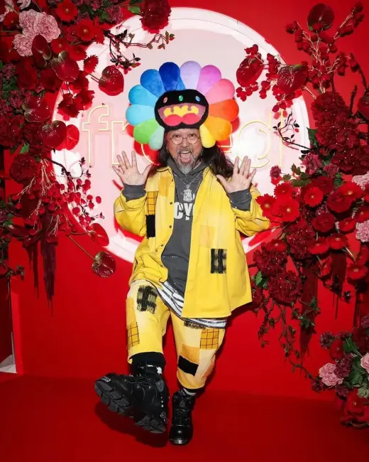 Takashi Murakami, rengarenk çiçek karakterli şapkası ve sarı kostümüyle kırmızı halıda poz veriyor. Sanatçının enerjik tarzı ve pop art estetiği dikkat çekiyor.