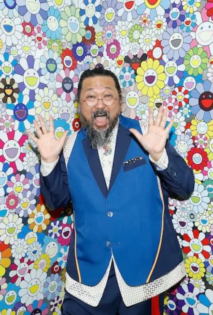 Takashi Murakami, kendine özgü çiçek desenleriyle kaplı renkli bir arka plan önünde enerjik bir poz veriyor. Sanatçının pop art ve neşeli görsel dili dikkat çekiyor.
