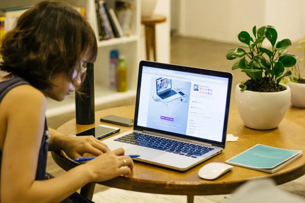 Evdeki çalışma ortamında online eğitim platformunda ders alan genç kadın; laptop, defter ve çiçek detaylarıyla sıcak bir öğrenme atmosferi.