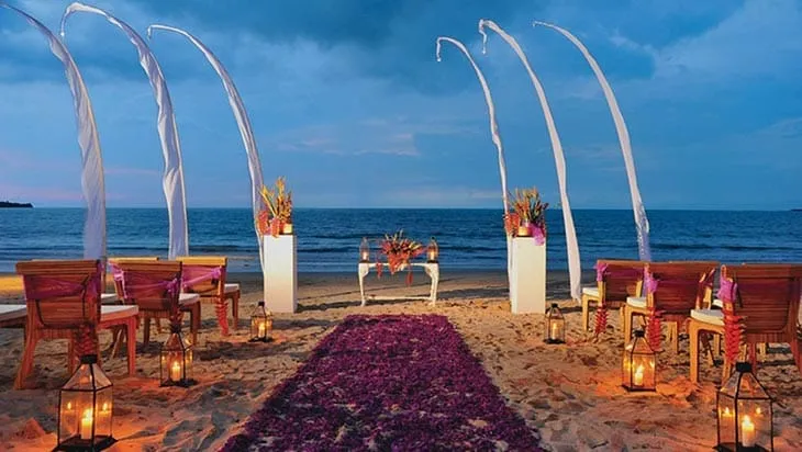 Bali sahilinde gün batımına karşı kurulmuş, mum ışıkları ve mor çiçeklerle süslenmiş romantik bir kumsal düğünü dekorasyonu.