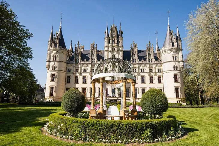Fransa’nın tarihi Château de Challain şatosunun bahçesinde yer alan, çiçeklerle süslenmiş ihtişamlı çardak ve arka planda masalsı şato manzarası.