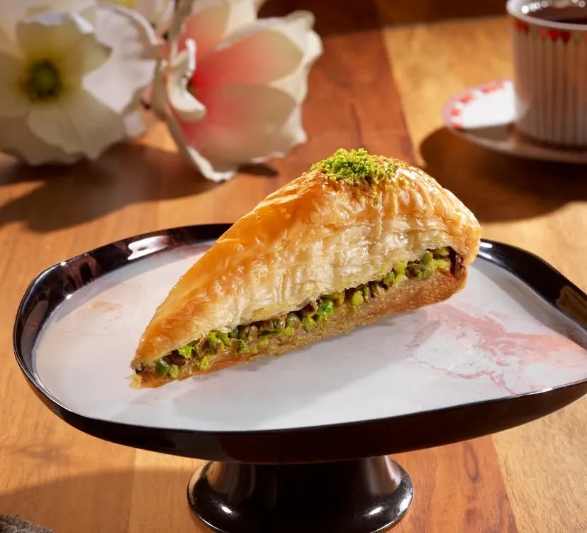 Türkiye'nin En İyi Baklavacıları rehberinde yer alan, incecik yufkalarla hazırlanan fıstıklı havuç dilimi baklava. Siyah ayaklı zarif beyaz bir tabakta sunulan bu çıtır geleneksel lezzete, ahşap masa üzerinde beyaz çiçekler ve ince belli bardakta taze demlenmiş Türk