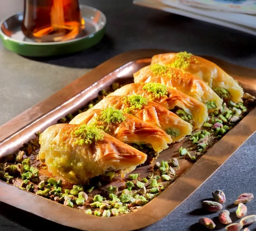 Bakır tepside sıralanmış, bol fıstık dolgulu ve üzeri Antep fıstığı tozuyla süslenmiş çıtır şöbiyet baklava dilimleri. Tepsi etrafına serpiştirilmiş fıstıklar ve arka planda ince belli bardakta geleneksel Türk çayı