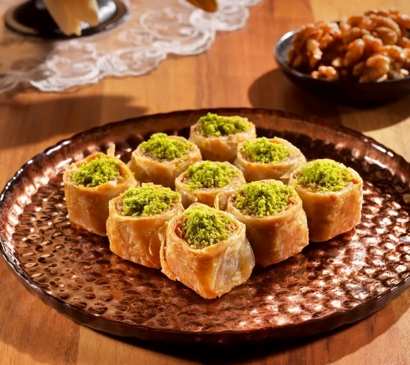 Dövme bakır tepside özenle dizilen, bol fıstıklı çıtır rulo baklavalar iştah açıcı biçimde sergileniyor. Arka plandaki ahşap masa, ceviz kasesi ve dantel detaylarıyla Türk mutfağının köklü geleneksel tatlı kültürü vurgulanıyor.