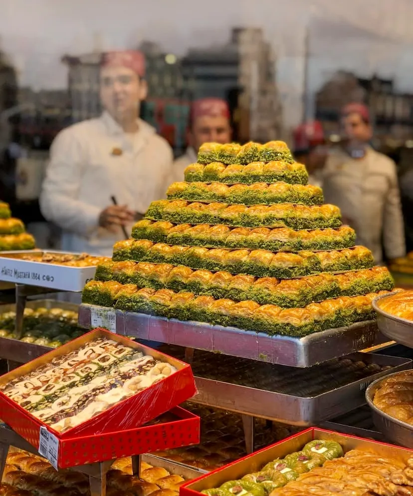 Türkiye’nin: Oggusto Türkiye'nin En İyi Baklavacıları: Vitrinde piramit formunda dizilmiş bol fıstıklı çıtır baklava tepsisi sergileniyor. Alt kısımdaki kırmızı kutularda lokum çeşitleri yer alırken, bulanık arka planda beyaz üniformalı, kırmızı şapkalı ustalar geleneksel tatlıları hazırlıyor.