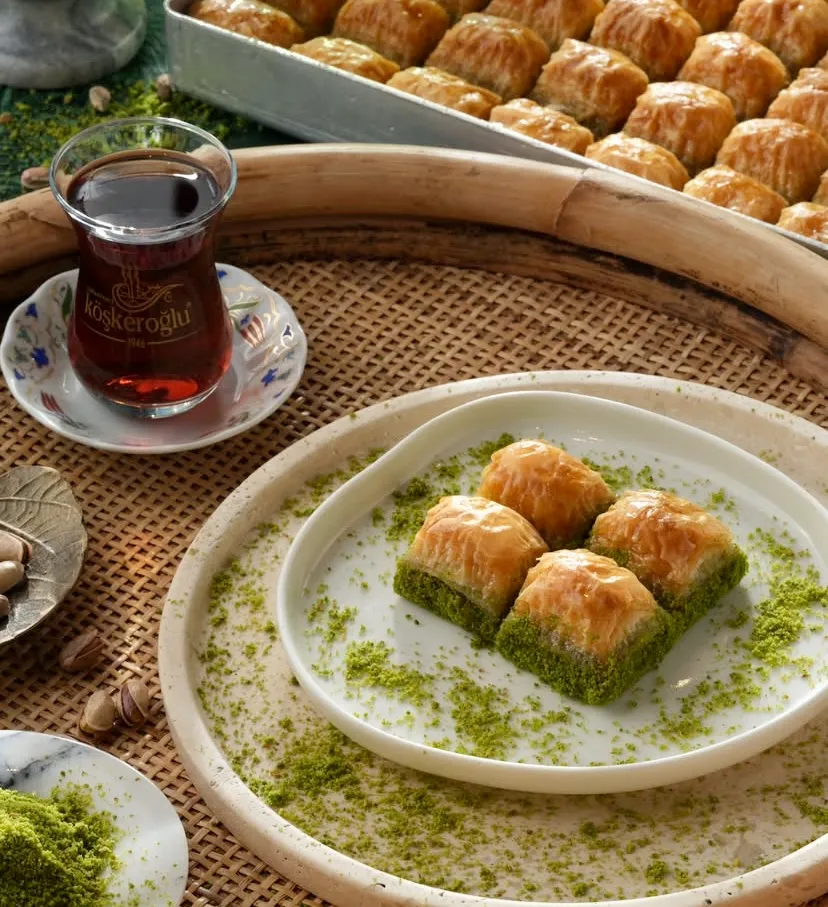 Köşkeroğlu logolu ince belli bardakta demli çay eşliğinde, beyaz tabakta sunulan dört dilim bol fıstıklı çıtır baklava. Hasır tepsiye serpiştirilmiş antep fıstığı tozları ve arka plandaki tepsi dolusu altın sarısı tatlılar iştah