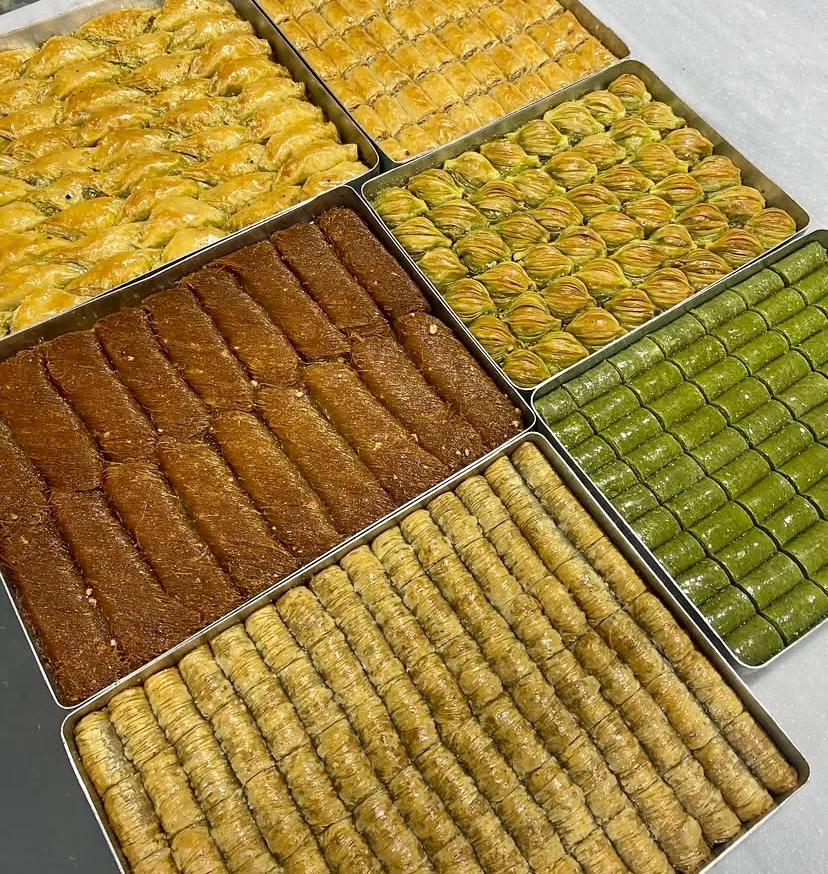 Altın sarısı ve fıstık yeşili tonlarındaki çıtır geleneksel tatlılar metal tepsilerde sergileniyor. Yan yana dizilmiş fıstıklı dürüm, midye baklava, şöbiyet ve burma kadayıf çeşitleri, Türk mutfağının eşsiz lezzet zenginliğini