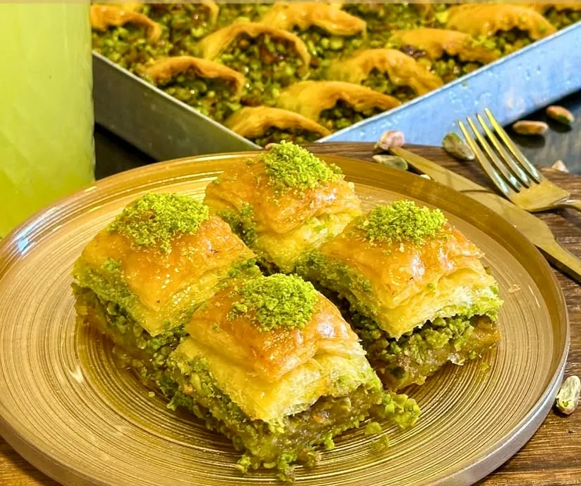 Altın sarısı tabakta sunulan, üzeri ve içi bol yeşil fıstık dolu dört dilim çıtır şerbetli baklava. Ahşap zeminli sunumun arka planında bir tepsi fıstıklı midye baklava, serinletici bir içecek ve şık çatal bıçak takımı yer
