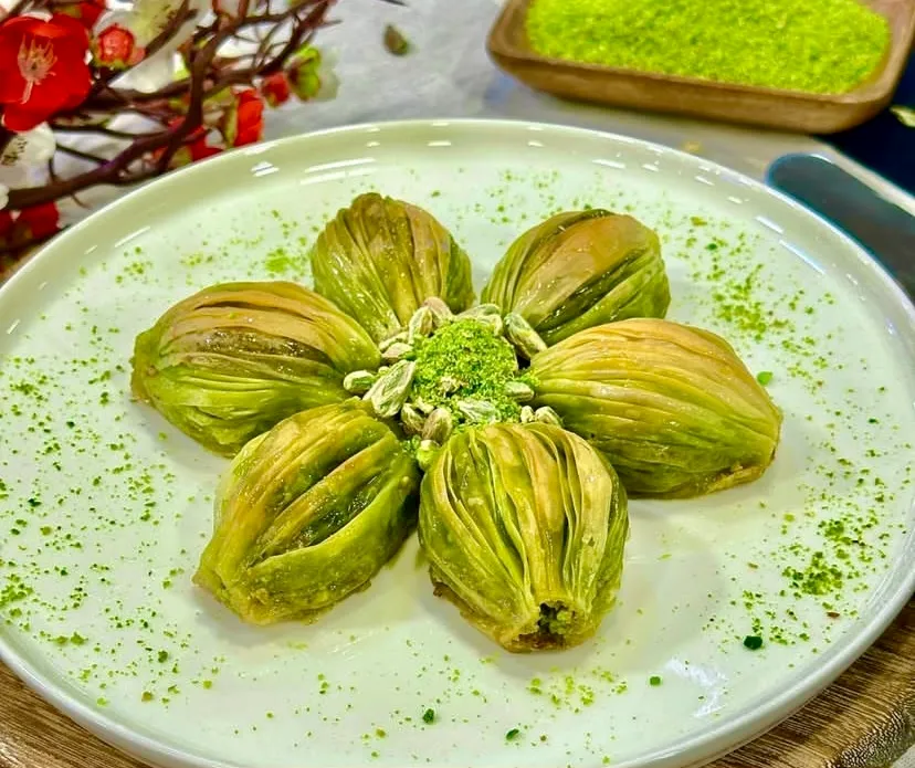 Beyaz tabakta çiçek formunda dizilmiş altı adet bol fıstıklı midye baklava. Merkezde Antep fıstığı tozu ve taneleri yer alırken, tabağın çevresine yeşil fıstık serpiştirilmiş. Arka planda kırmızı çiçekler ve fıstık dolu ahşap kaseyle