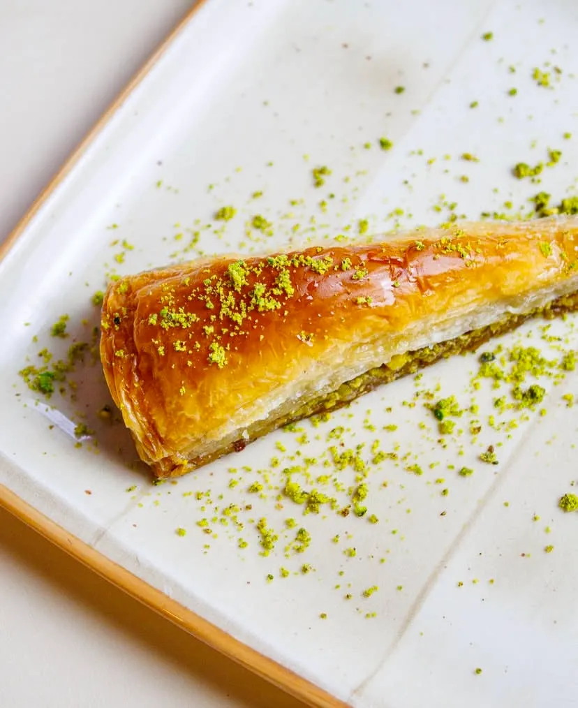 Beyaz seramik tabakta sunulan, ince açılmış yufkasıyla altın sarısı çıtır havuç dilimi baklava. Tatlının arasındaki kalın Antep fıstığı dolgusu öne çıkarken, parlak yüzeyine ve tabağın geneline ince çekilmiş yeşil fıstık tozu