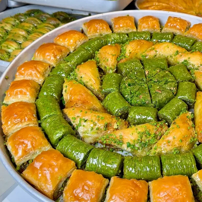 Yuvarlak metal tepsiye dairesel düzenle dizilmiş çeşitli şerbetli tatlılar. Dış halkada altın sarısı çıtır kare baklavalar, içe doğru parlak yeşil fıstıklı dürümler, üçgen şöbiyetler ve merkezde yoğun fıstıklı özel kesimler