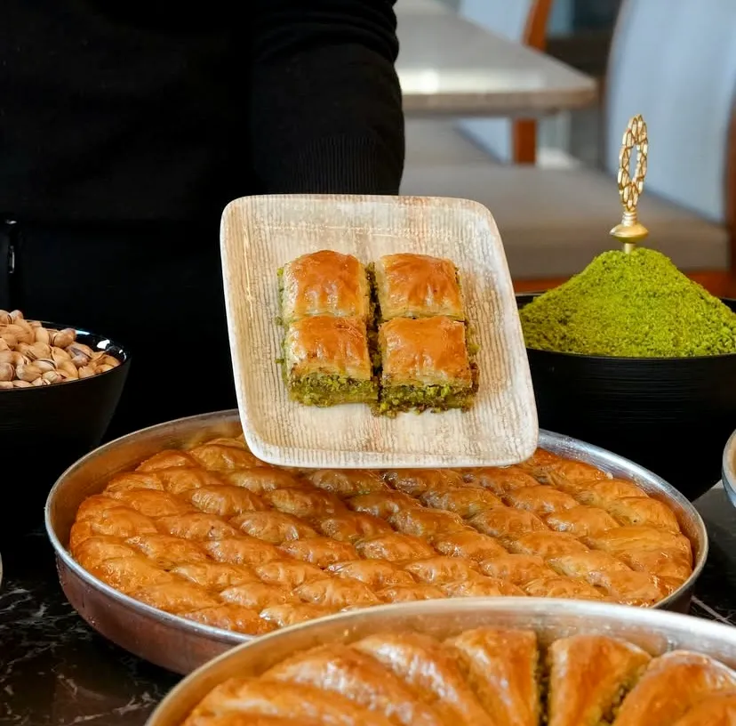 Siyah giyimli biri kare tabakta dört dilim fıstıklı çıtır baklava sunuyor. Alt kısımda metal tepsilerde nar gibi kızarmış şerbetli baklavalar, yanlarda ise taze bütün ve ince çekilmiş toz Antep fıstığı dolu siyah kaseler sergileniyor.