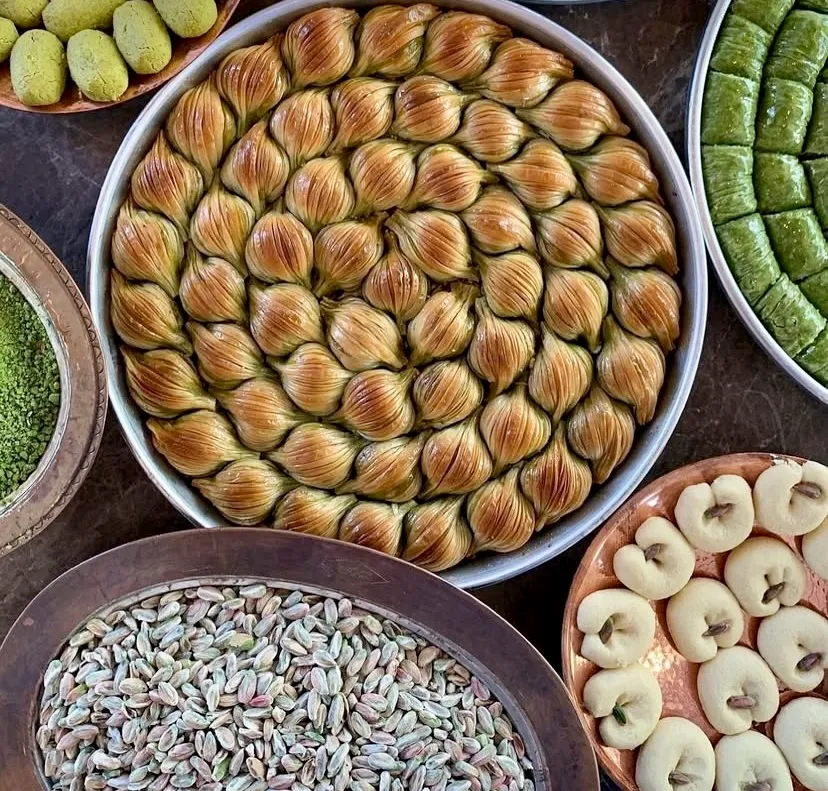 Geleneksel yuvarlak tepside sarmal düzende dizilmiş çıtır midye baklavalar; etrafını saran taze iç fıstık dolu bakır kâseler, fıstıklı dürümler ve kurabiyeler eşliğinde Türk mutfağının köklü tatlı zanaatını kusursuzca sergiliyor.