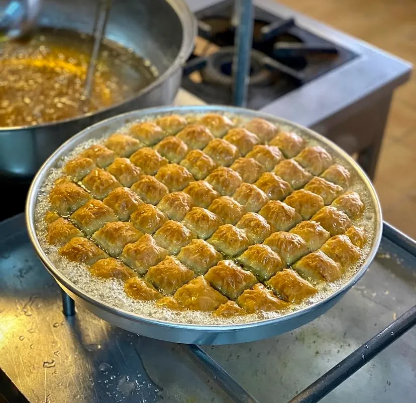 Türkiye’nin: Oggusto Türkiye'nin En İyi Baklavacıları: Profesyonel mutfak ocağında, yuvarlak metal tepsi içindeki taze pişmiş altın sarısı baklava dilimleri üzerinde köpüren sıcak şerbet. Arka plandaki kaynayan şerbet tenceresiyle, ustalık gerektiren geleneksel tatlı yapım aşaması
