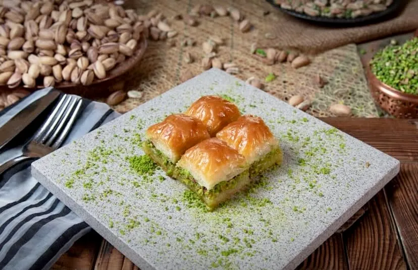 Türkiye’nin: Oggusto, Türkiye'nin En İyi Baklavacıları: Ahşap masada gri taş tabakta sunulan, yeşil fıstık tozuyla süslenmiş dört dilim altın sarısı çıtır fıstıklı baklava. Arka plandaki taze Antep fıstıklarıyla zenginleşen bu sunum, Türk mutfağının ipek inceliğindeki köklü tatlı geleneğini
