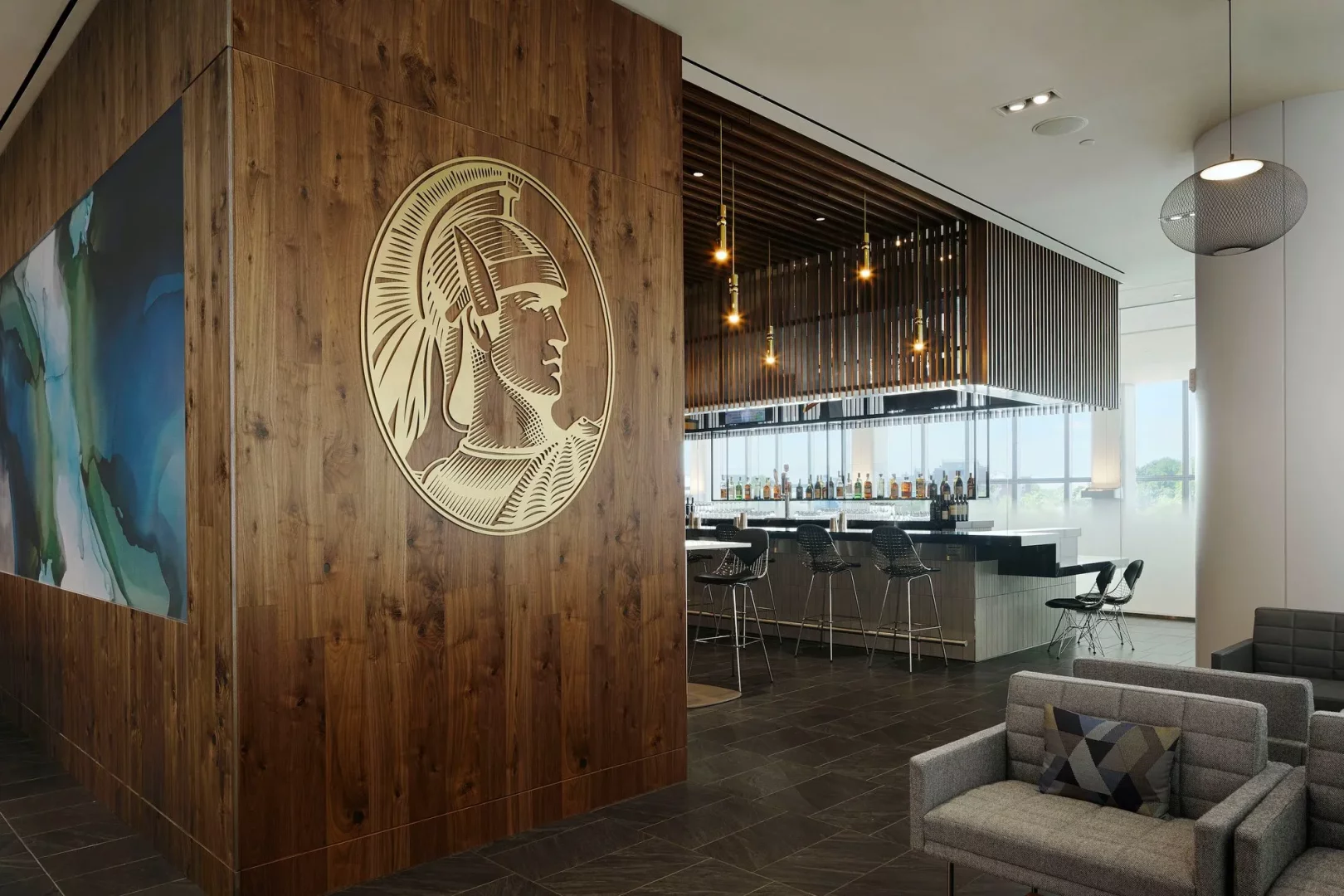 Ahşap duvarında büyük Amex Centurion logosu ve soyut mavi-yeşil bir sanat eseri bulunan, modern tasarımlı bu havalimanı lounge'unda, şık bir bar alanı ve gri koltuklar lüks bir seyahat öncesi dinlenme ortamı sunuyor.