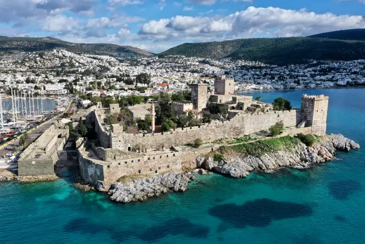 Masalsı mavi sularla çevrili, kayalık bir yarımada üzerinde görkemli bir şekilde yükselen Bodrum Kalesi, tarihi surları, kuleleri ve içindeki cami minaresiyle öne çıkarken, arkasında modern Bodrum şehrinin beyaz evleri ve canlı marinası uzanıyor.