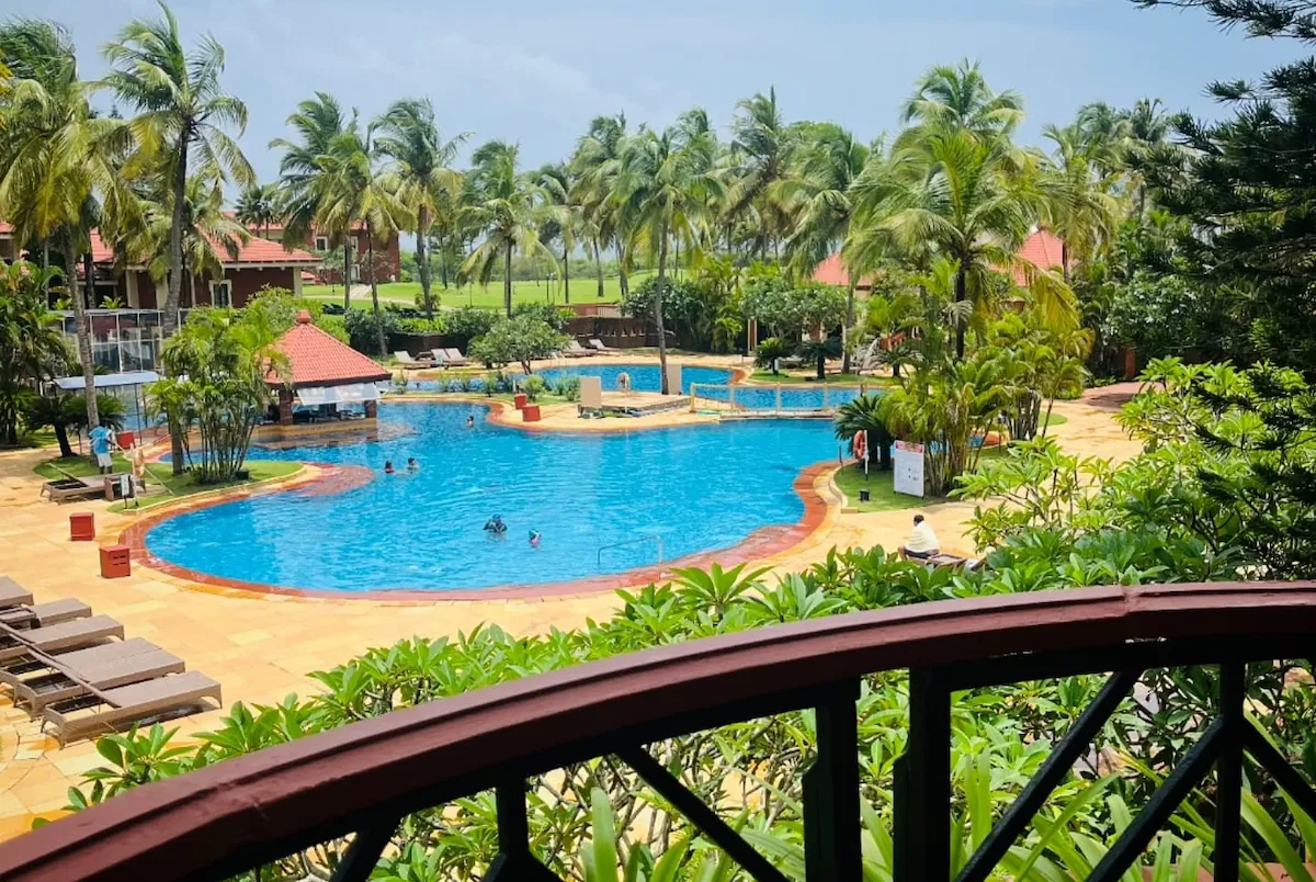 Caravela Beach Resort'un yemyeşil palmiye ağaçları ve tropikal bitki örtüsüyle çevrili, içinde yüzen insanların olduğu, kırmızı çatılı bir havuz barı da bulunan geniş mavi yüzme havuzu kompleksinin, uzaktan görünen golf sahasıyla birlikte bir balkondan çekilmiş kuşbakışı görüntüsü.