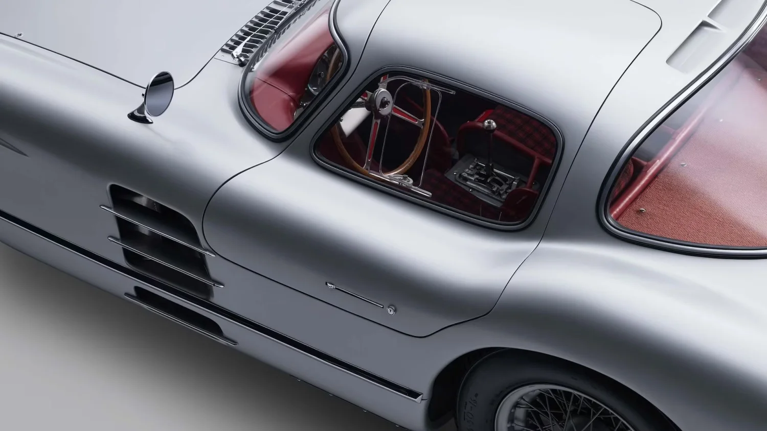 Dünyanın en pahalı otomobili unvanına sahip gümüş rengi Mercedes 300 SLR Uhlenhaut Coupe'nin yan profilini gösteren bu yakın çekim fotoğrafta, aracın göz alıcı kırmızı ekose iç döşemesi, zarif ahşap direksiyonu ve krom detaylarıyla klasik tasarımı öne çıkmaktadır.