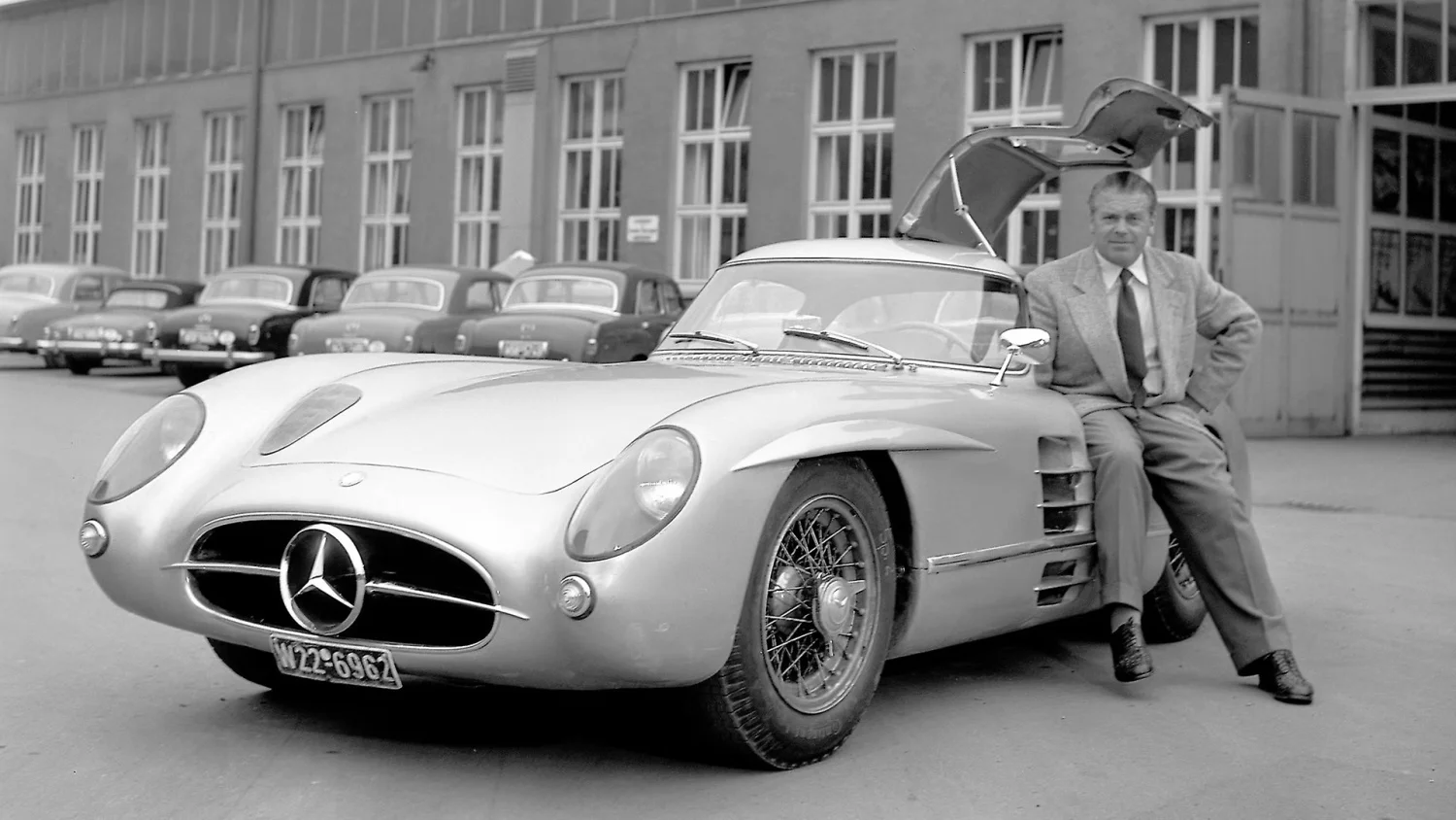 Siyah beyaz bir fotoğrafta, martı kanadı kapısı yukarı doğru açık duran, zarif gümüş rengi Mercedes-Benz 300 SLR model otomobilin yanına Rudolf Uhlenhaut oturmuş, bu ikonik aracın dünyanın en pahalı otomobili olarak ününe vurgu yapıyor ve arkasında diğer klasik araçlar sıralanıyor.