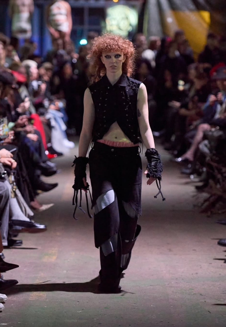 Kopenhag Moda Haftası Sonbahar/Kış 2025 defilesinde, kıvırcık kızıl saçları ve belirgin makyajıyla dikkat çeken bir model, Fine Chaos markasının ham dokular ve çarpıcı kontrastlarla bezeli özgün koleksiyonundan, asimetrik kesimli kısa siyah yelek, gümüş rengi yamalı bol pantolon ve püsküllü eldiven benzeri aksesuarlardan oluşan iddialı bir kombinle podyumda yürüyor.
