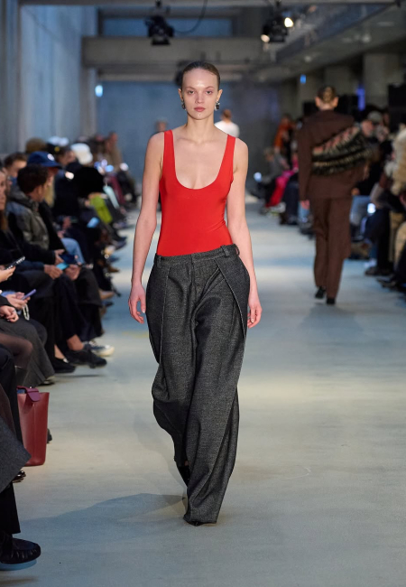 Forza Collective'in Sonbahar/Kış 2025 Kopenhag Moda Haftası defilesinde, canlı kırmızı, U yakalı bir atlet üst ve önünde belirgin dikiş detayları olan, hacimli, koyu gri bol paça bir pantolon giyen bir model, iyi aydınlatılmış podyumda izleyicilerin önünden yürüyor.