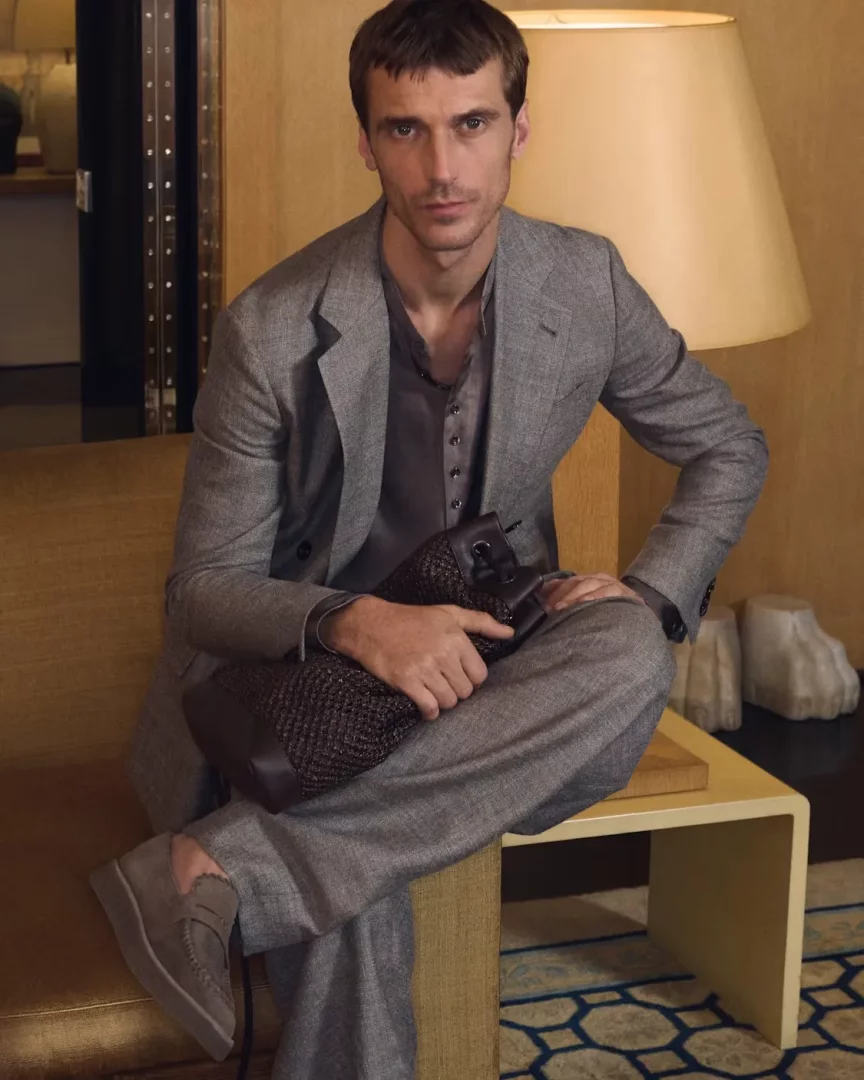 Giorgio Armani tarzında, rahat kesimli gri bir takım elbise ve süet loafer ayakkabılar giyen, çapraz bacak oturan ve dokulu koyu renk bir çanta tutan genç bir adam, arkasında sıcak tonlu duvarlar ve büyük bir lambanın bulunduğu şık bir odada ciddi ifadeyle kameraya bakıyor.