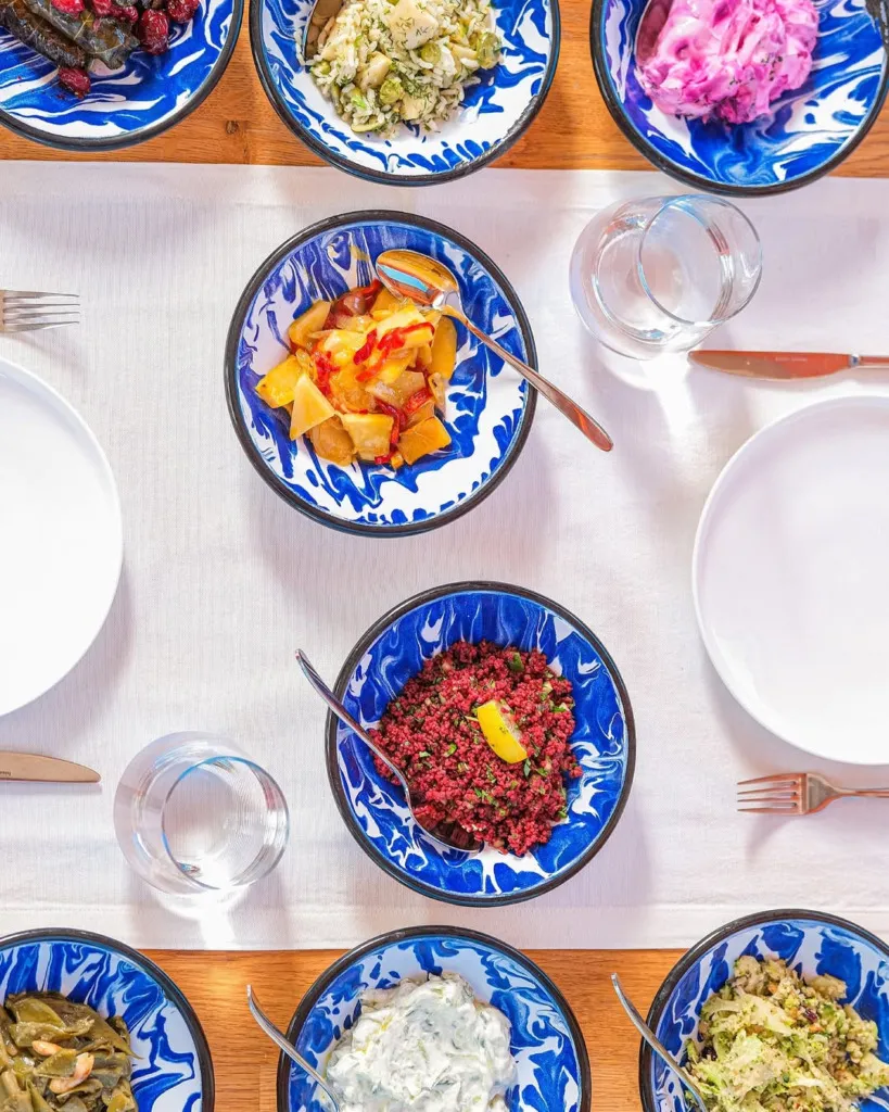 Beyaz masa örtüsü serili ahşap bir masa üzerinde, Bodrum mutfağının zenginliğini yansıtan, mavili beyazlı desenli kaselerde sunulan zeytinyağlı yaprak sarma, pancar salatası, kısır, patates, deniz börülcesi gibi çeşitli meze tabakları ve yanlarında boş servis tabakları ile su kadehleri dolu bir ziyafet masası kuşbakışı görünüyor.