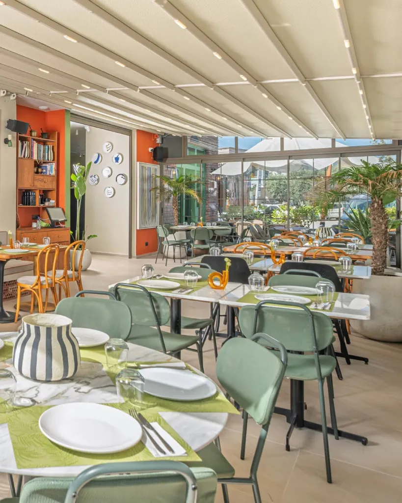 Bodrum'daki Havandan restoranının aydınlık iç mekanında, yeşil metal sandalyelerle çevrili mermer masalar yeşil suplalarla donatılmış, açılır kapanır pergola tavan doğal ışık sağlarken cam pencerelerden dışarıdaki yeşil bitkiler görülüyor.