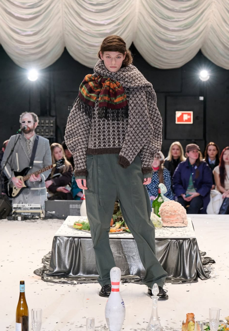 Henrik Vibskov'un Sonbahar/Kış 2025 Kopenhag Moda Haftası defilesinde, örgü desenli, hacimli bir kazak, üzerine renkli püsküllü bir şal ve bol paça koyu yeşil bir pantolon giyen model, retro esintili bu kat kat kombini sunarken arkada canlı müzik performansı ve seyirciler yer alıyor.