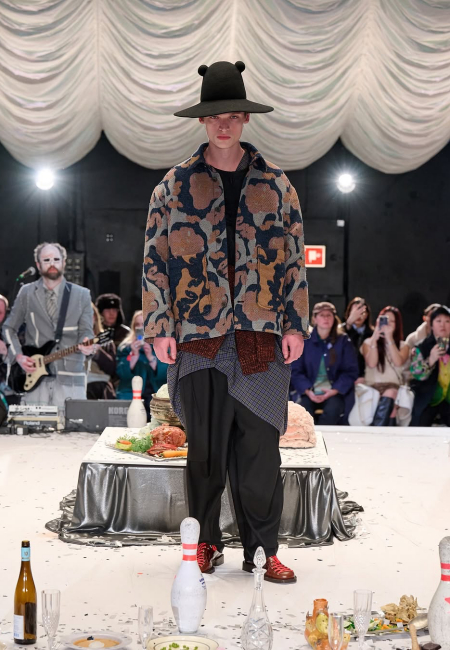 Henrik Vibskov'un Sonbahar/Kış 2025 Kopenhag Moda Haftası defilesinde, kulak detaylı şapka, desenli katmanlı ceket, asimetrik ekose gömlek ve bol pantolondan oluşan retro esintili bir kombinle podyumda yürüyen model görülüyor.