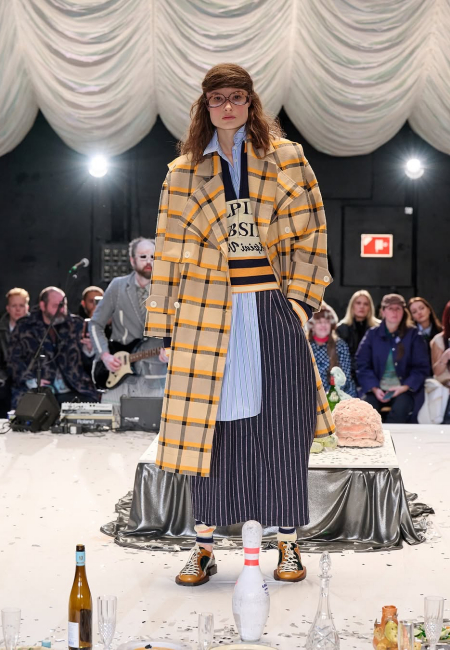 Sonbahar/Kış 2025 Kopenhag Moda Haftası defilelerinde Henrik Vibskov'un katmanlı ve retro esintili koleksiyonundan bir anı gösteren bu karede, ekose trençkotu, çizgili süveteri ve eteğiyle dikkat çeken kahverengi çerçeveli gözlüklü bir model, konfeti dolu bir podyumda, arka planda müzik çalan bir grubun önünde yürüyor.