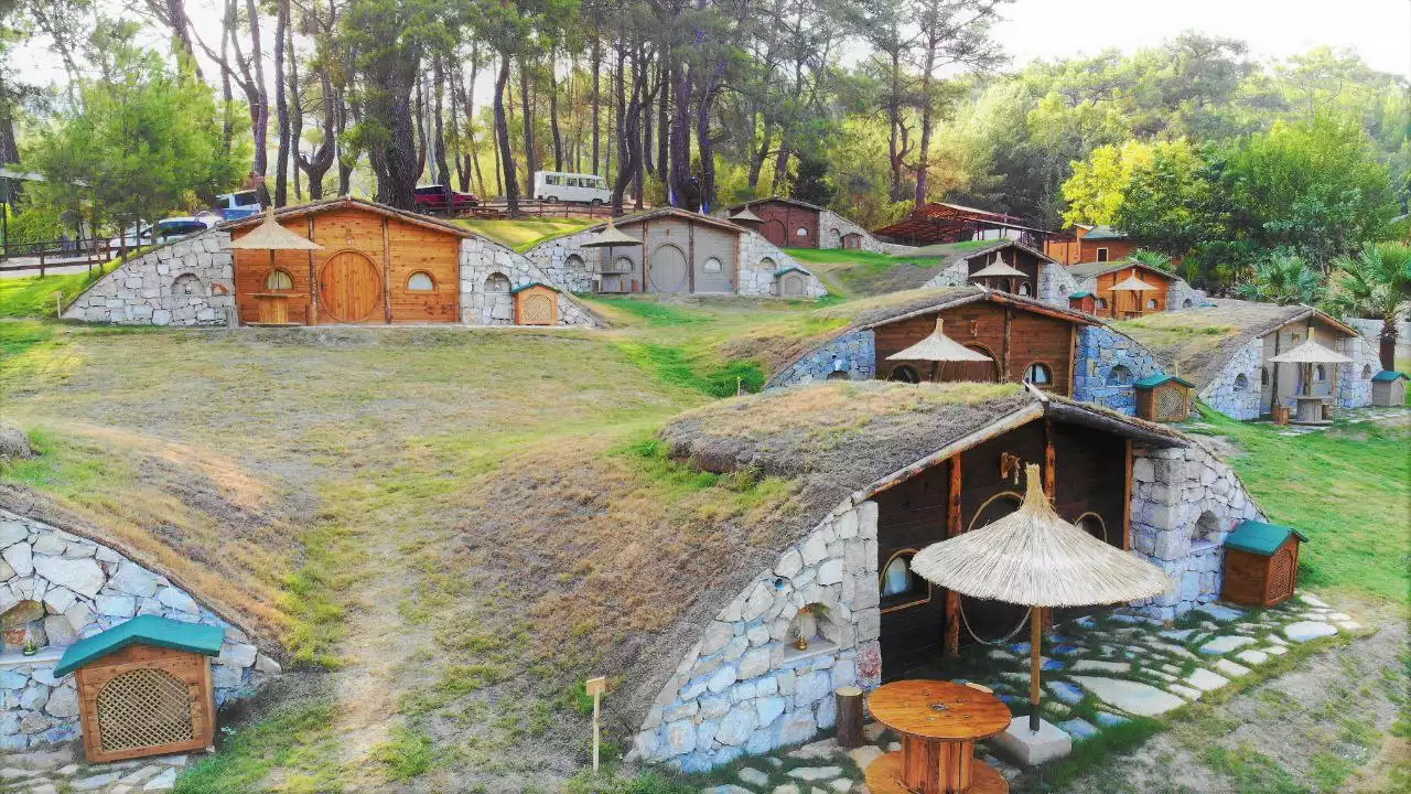 Ormanlık bir alanda, yeşil bir yamaca gömülü Hobbit evleri. Taş temelleri, yuvarlak ahşap kapıları ve çim çatılarıyla fantastik bir konaklama köyü atmosferi yaratıyor.