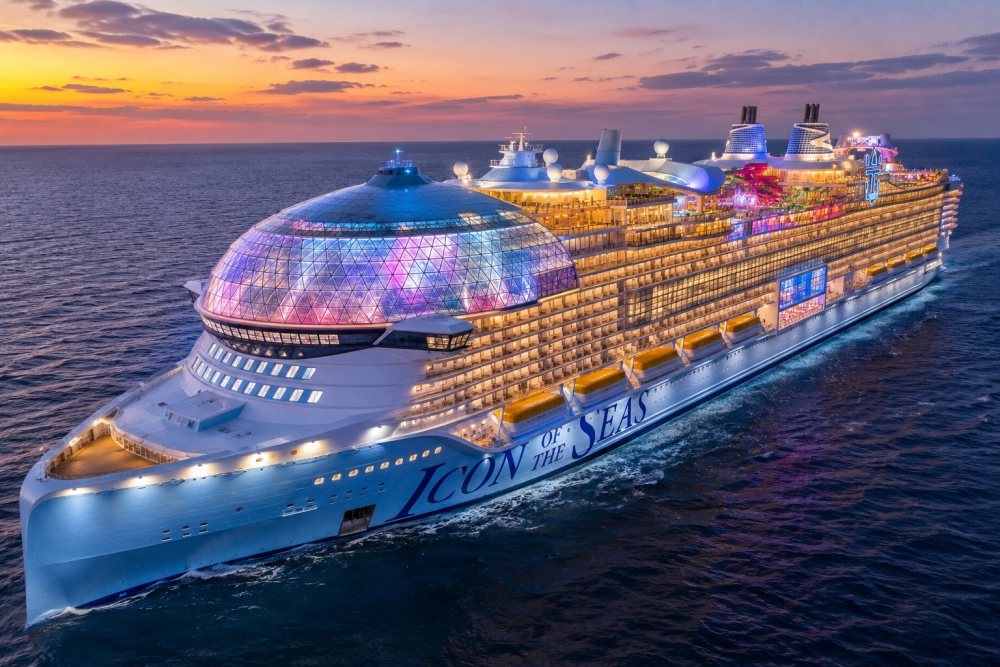 Denizde ilerleyen, birçok ışıkla aydınlatılmış ultra lüks Royal Caribbean Icon of the Seas kruvaziyer gemisi, gün batımında veya alacakaranlıkta, ön güvertesindeki çok renkli ışıklarla parlayan büyük cam kubbesiyle dikkat çekiyor.