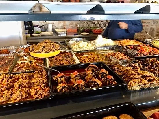 Çeşit çeşit, taze hazırlanmış ev yemeklerinin sergilendiği, pilav, köfte, güveç ve dolma gibi geleneksel lezzetlerle dolu bu büfe hattında, arkada mavi gömlekli bir çalışan müşterilere sıcak servis yapmaya hazırlanırken, sunulan her bir tabak adeta iki lokmada doyurucu bir ziyafet vaat ediyor.