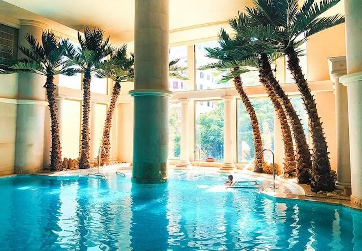 Beyrut'taki InterContinental Phoenicia otelinin lüks iç mekan havuzunda, çevresinde sıralanmış uzun palmiye ağaçları ve dışarıyı gösteren geniş pencerelerle tropikal bir atmosfer sunulurken, suda keyifli vakit geçiren bir misafir görülüyor.