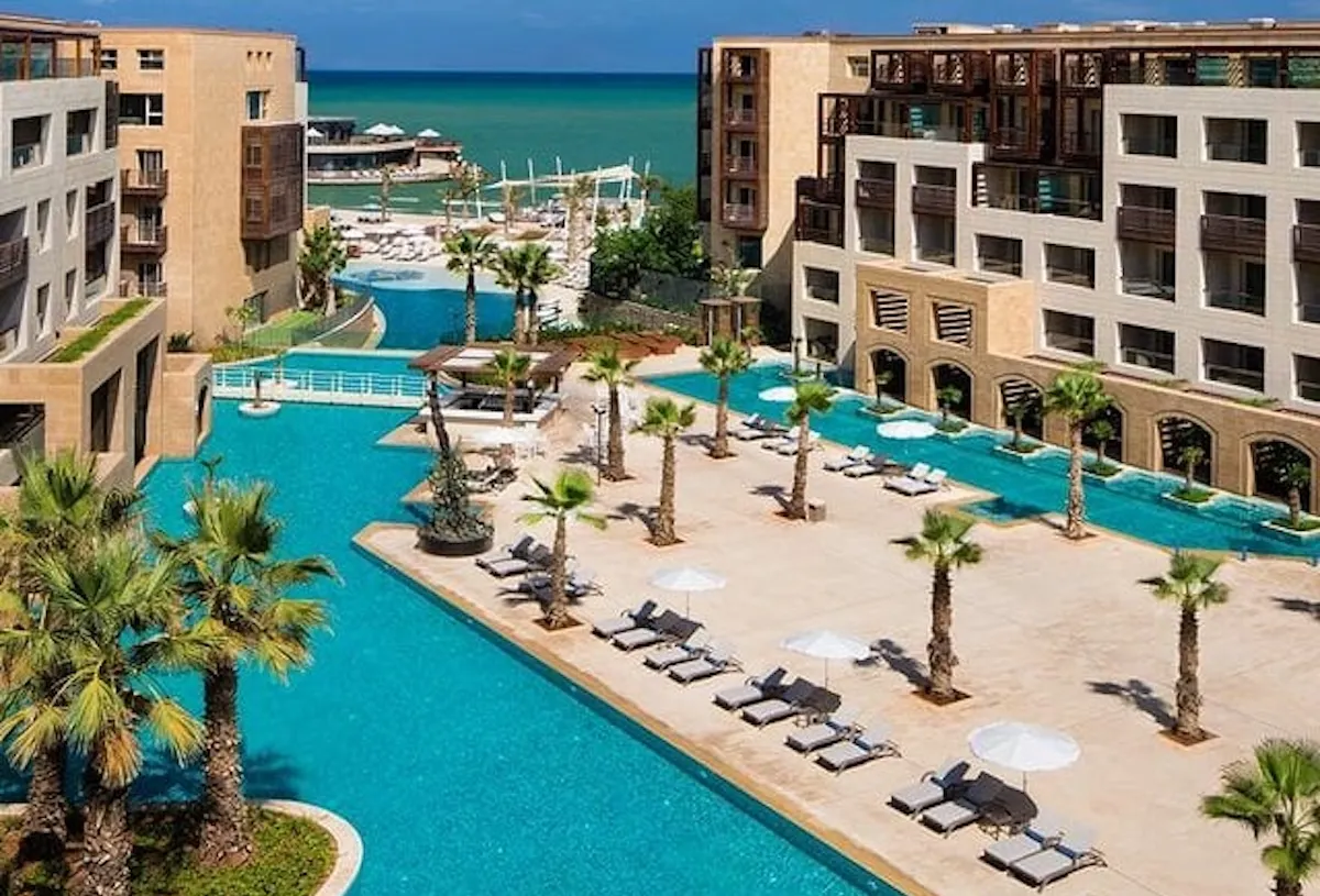 Kempinski Summerland Beyrut tesisinde, Akdeniz'e doğru uzanan, palmiye ağaçları, beyaz şezlonglar ve şemsiyelerle çevrili berrak turkuaz havuzların kuşbakışı görüntüsü, rahatlatıcı bir yaz tatili atmosferi sunan lüks bir kompleks gösteriyor.