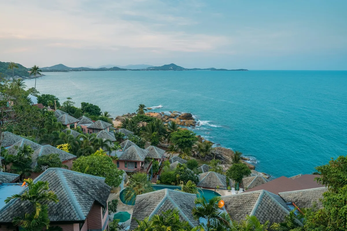 Yukarıdan çekilmiş bu geniş açılı fotoğrafta, yemyeşil tropik bitki örtüsüyle çevrili, kiremit çatılı lüks villalar ve bazılarına ait özel havuzlar Koh Samui'nin masmavi deniziyle buluşan kayalık kıyısı boyunca uzanıyor, uzakta adalar belirginleşerek huzurlu bir tatil manzarası oluşturuyor; en güzel balayı rotaları için ilham veren bir kare.