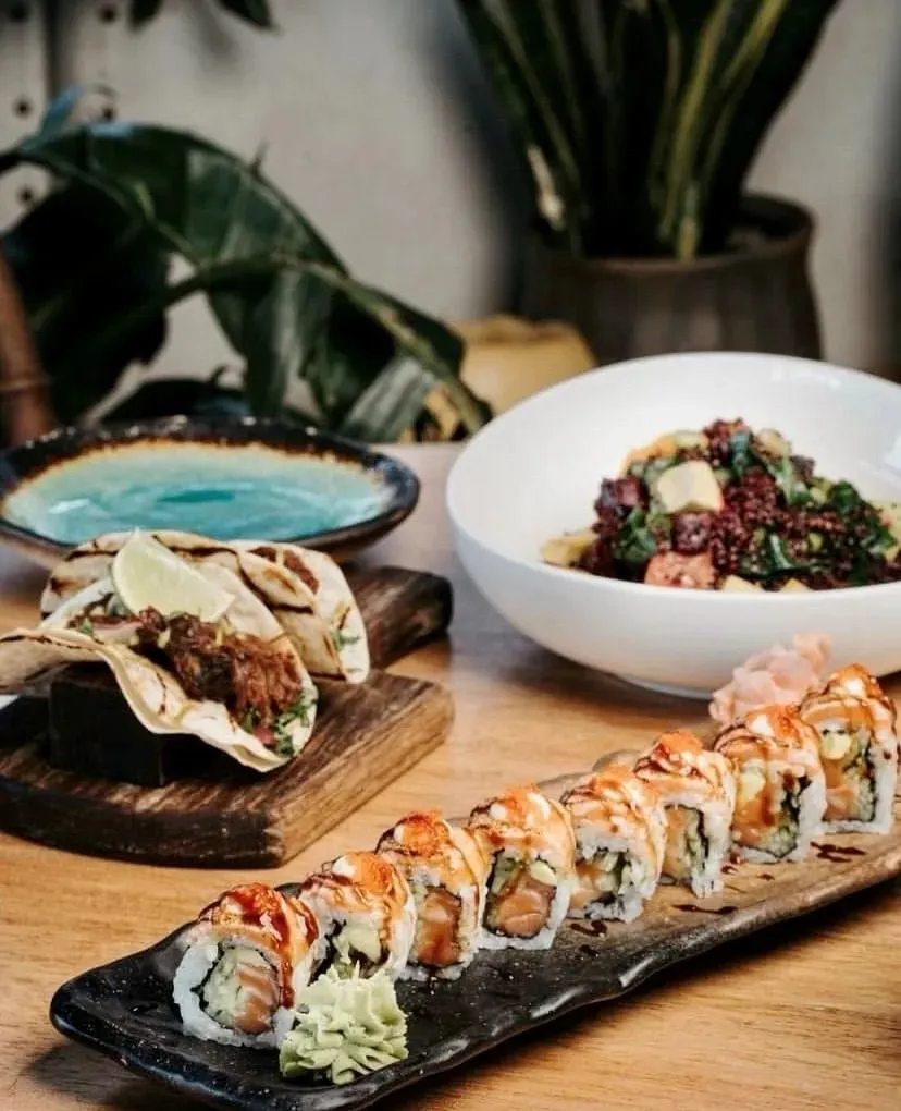 Tahta bir masada servis edilen sushi ruloları, taco ve renkli bir salata tabağı.