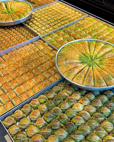 Oggusto Türkiye'nin En İyi Baklavacıları rehberindeki geleneksel lezzetleri yansıtan tepsiler: Altın sarısı çıtır yufkalı klasik kare baklavalar, yuvarlak tepside bol fıstıklı üçgen havuç dilimi ve alt sırada içi fıstık dolgulu yeşil-sarı renkli midye baklavalar yan yana