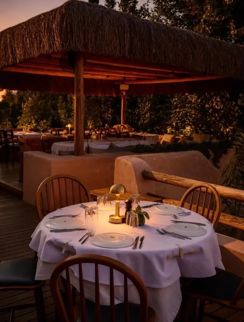 Bitez'deki Kurul restoranının açık hava bölümünde, hasır çatılı bir alanda, beyaz masa örtülü yuvarlak masalar, tabaklar, çatal bıçaklar ve ortalarında sıcak bir ışık yayan masa lambalarıyla akşam yemeği için özenle hazırlanmış samimi ve zarif bir ortam.