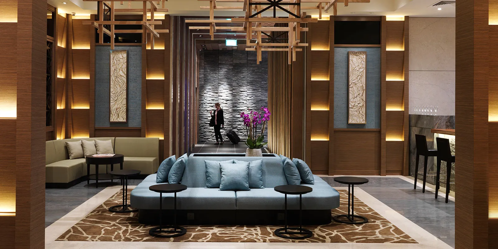 Lüks ve modern tasarımlı bir havalimanı lounge'unun iç görünümü; ortada açık mavi minderli uzun bir koltuk, solunda bej L şekilli kanepe, sağda bar alanı ve arka planda mor orkidelerin yer aldığı, bavuluyla yürüyen bir yolcunun görüldüğü taş duvarlı koridor, ahşap panelli duvarlardaki gizli şerit aydınlatmalarla sıcak bir atmosfer sunarak üst düzey seyahat konforunu vurguluyor.