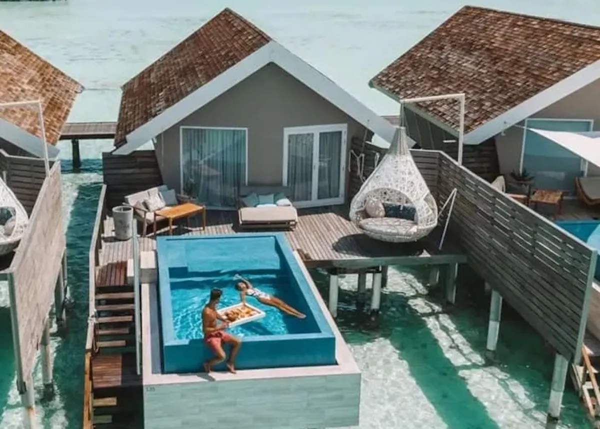 Maldivler'deki lüks LUX South Ari Atoll resortunda, havuzlu bir overwater villanın özel güvertesinde yüzen bir kahvaltı tepsisinin yanındaki havuzda yüzen bir kadın ve havuz kenarında oturan bir erkek, muhteşem turkuaz deniz manzarası eşliğinde tatilin keyfini çıkarıyor; en güzel balayı rotaları için ilham veren bir kare.
