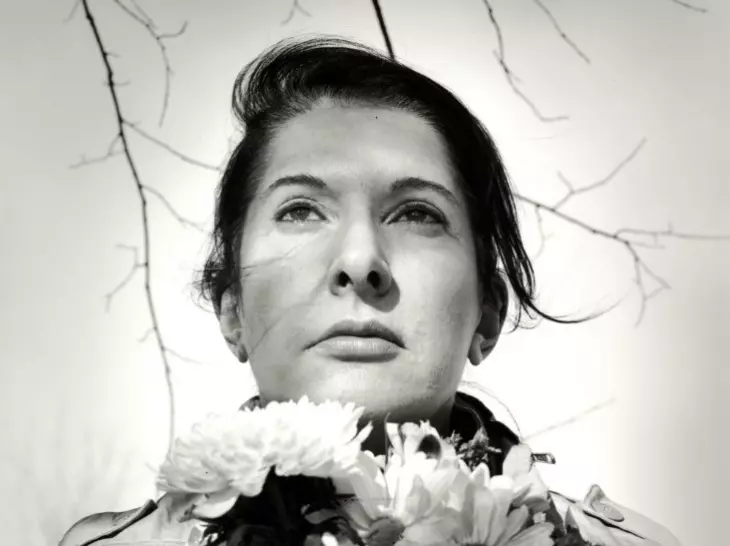Marina Abramović’in siyah beyaz portresi; sanatçı yüzünde kararlı bir ifade ve boynunda çiçeklerle, performans sanatındaki ruhsal derinliğini yansıtıyor.