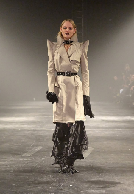 Kopenhag Moda Haftası'nda bir defilede, güçlü omuz detaylarına sahip bej trençkotu, siyah kemeri, boğazındaki kayışlı aksesuarı ve volümlü koyu renk alt giysisiyle dikkat çeken bir model, markanın feminen ve maskülen öğeleri harmanlayan Sonbahar/Kış 2025 koleksiyonundan bir tasarımı sergiliyor.